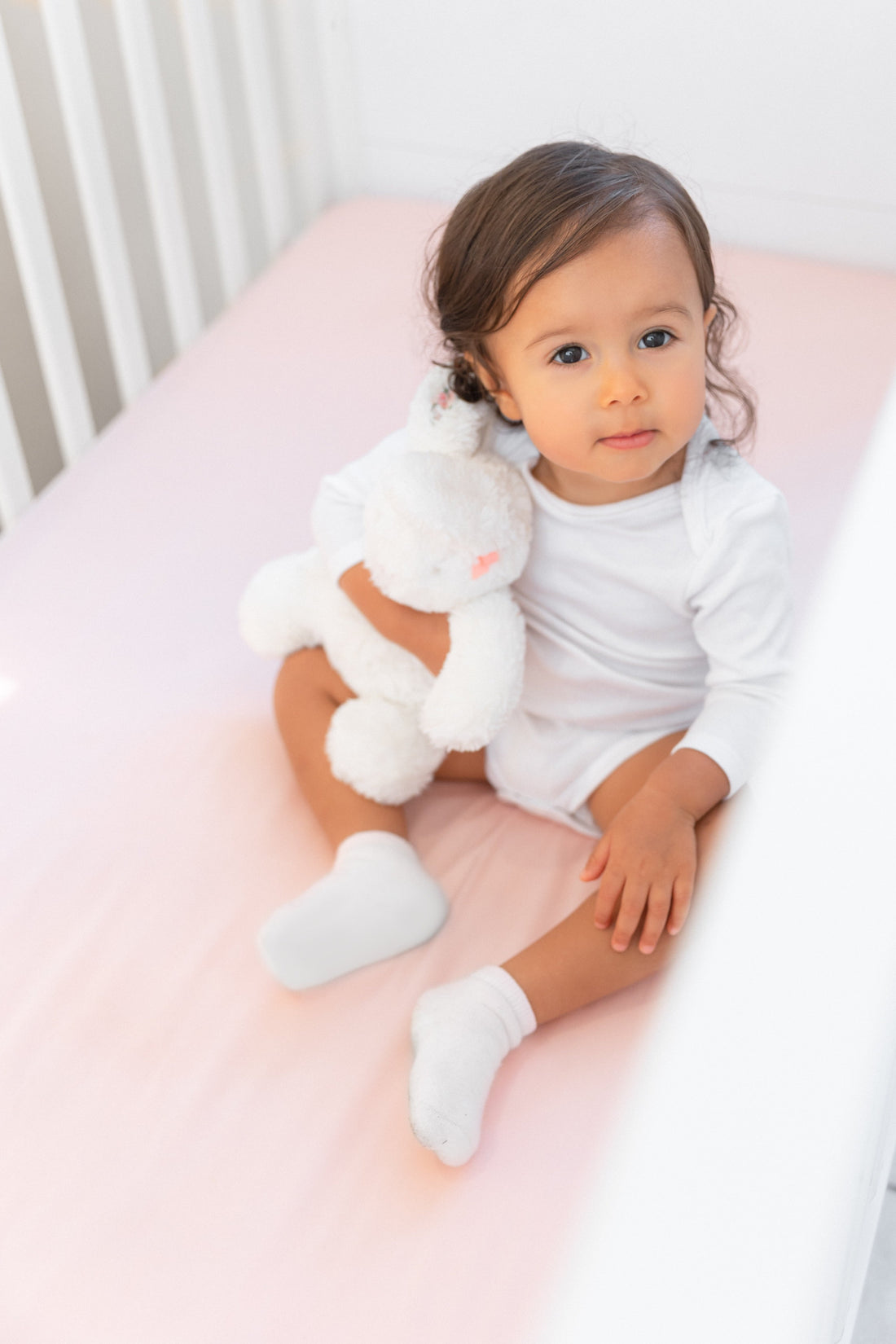 beautiful ittle girl with soft toy sitting in crib with BeddyByes Blush Pink Silk Cotbed Fitted Sheet