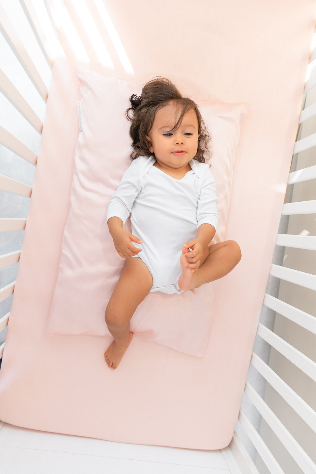 Toddler girl laying on a BeddyByes Blush Pink Silk Cotbed Fitted Sheet